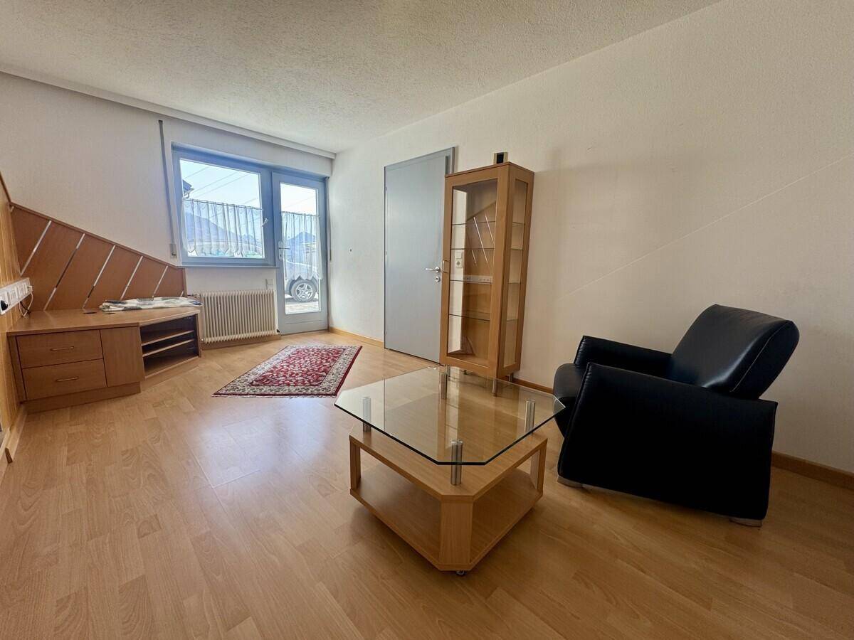Heller Raum mit Holzboden, Sessel und Glasvitrine, mit Zugang zu einem Balkon.