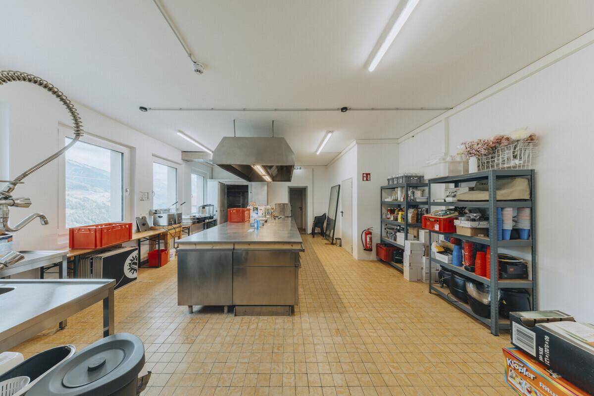Geräumige Industrieküche mit Edelstahlmöbeln, großer Dunstabzugshaube und gefliestem Boden, ideal für professionelles Kochen.