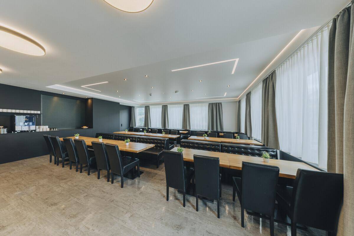 Geräumiger Speisesaal mit modernen Holztischen, schwarzen Stühlen und gepolsterten Sitzbänken, beleuchtet von großen Fenstern.