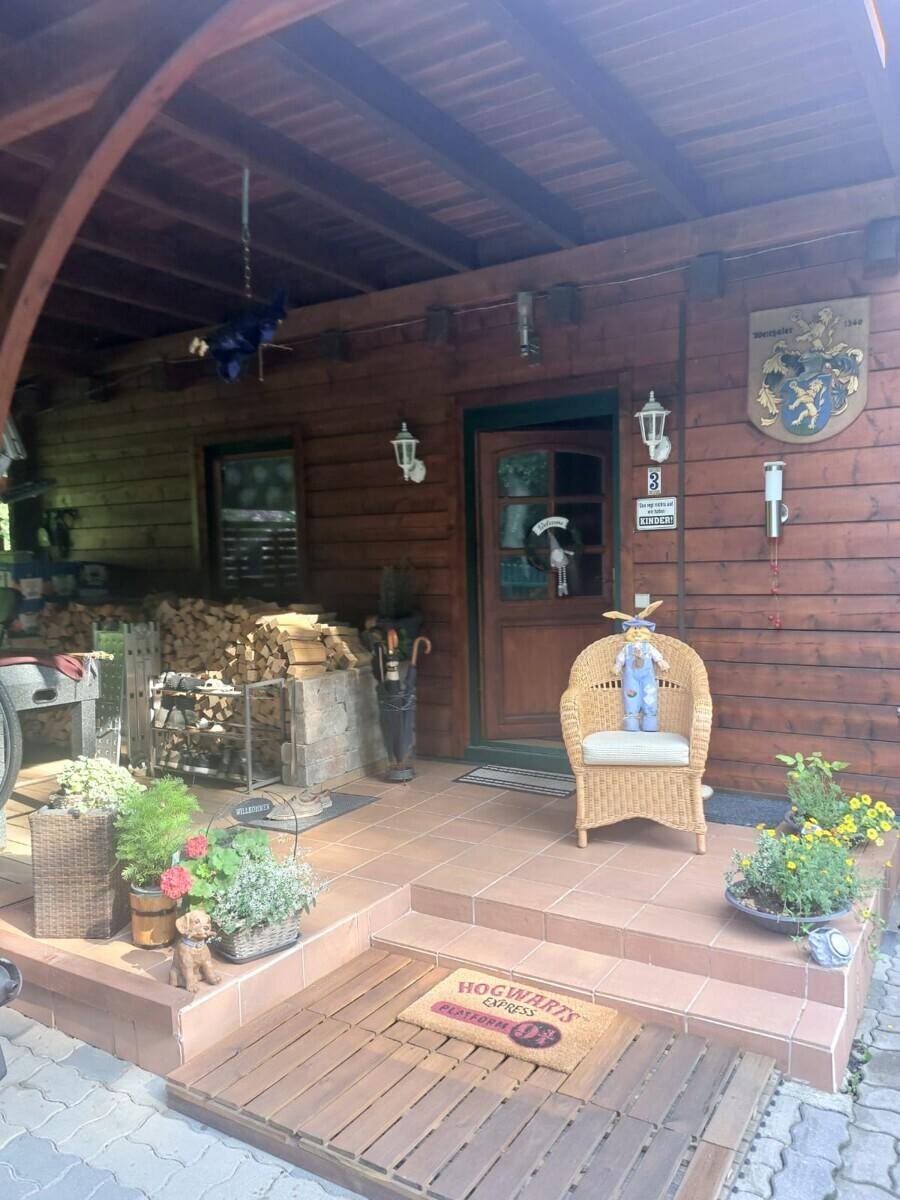 Überdachter Eingangsbereich des Holzhauses mit Korbsessel, Pflanzen und rustikaler Dekoration.