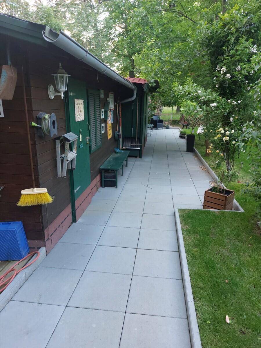 Gepflasterter Gehweg entlang des Holzhauses, umgeben von Bäumen und Sträuchern im Garten.