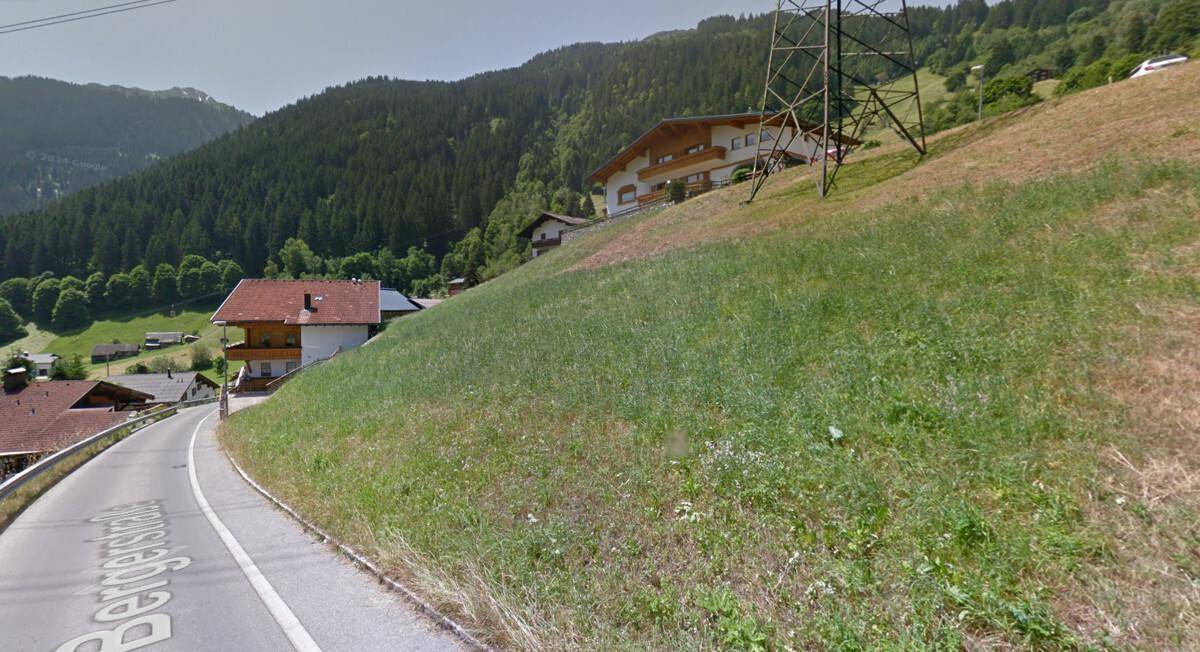 Blick auf eine grüne Wiese und eine Straße, die sich einen Hang hinaufwindet, mit Häusern und Bergen im Hintergrund.
