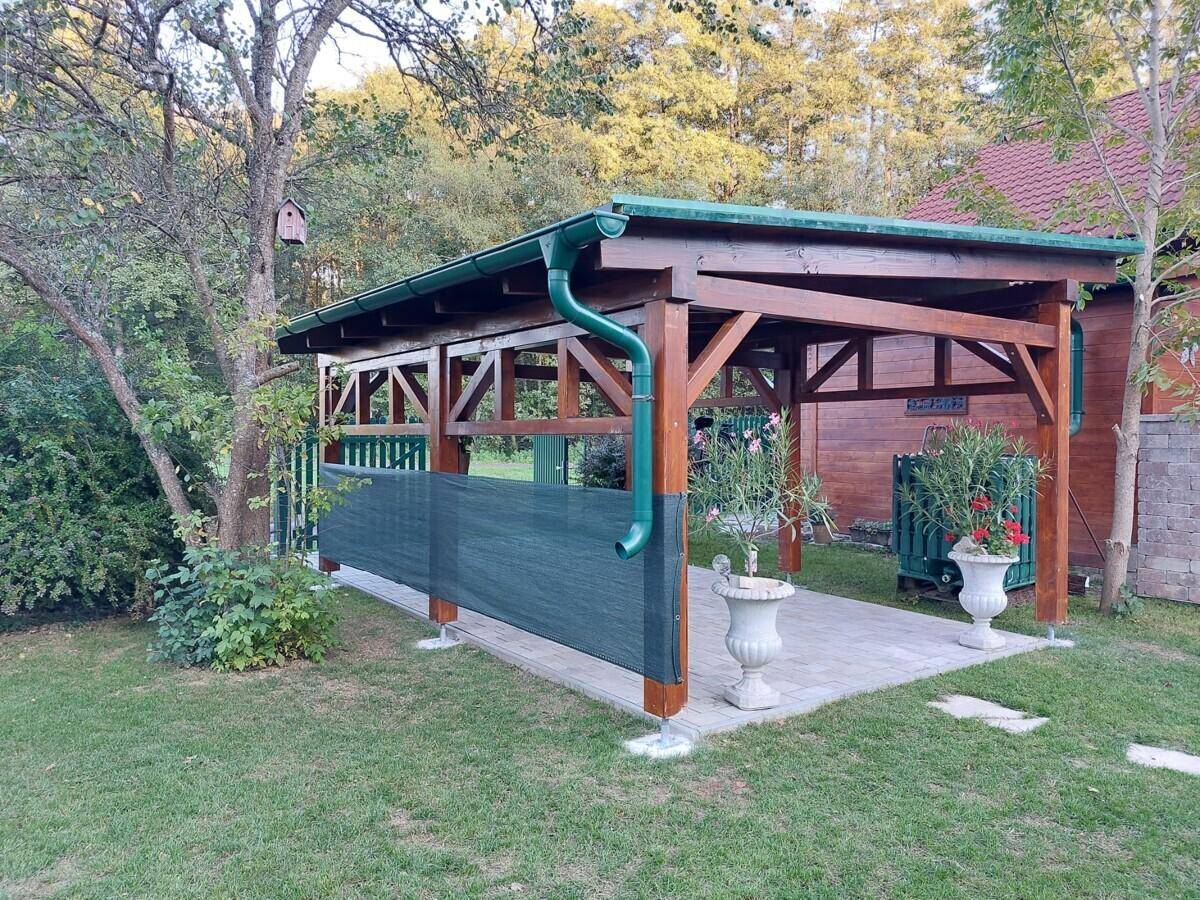 Freistehende Holzpergola mit gepflastertem Bereich im Garten, umgeben von Bäumen und Grünflächen.
