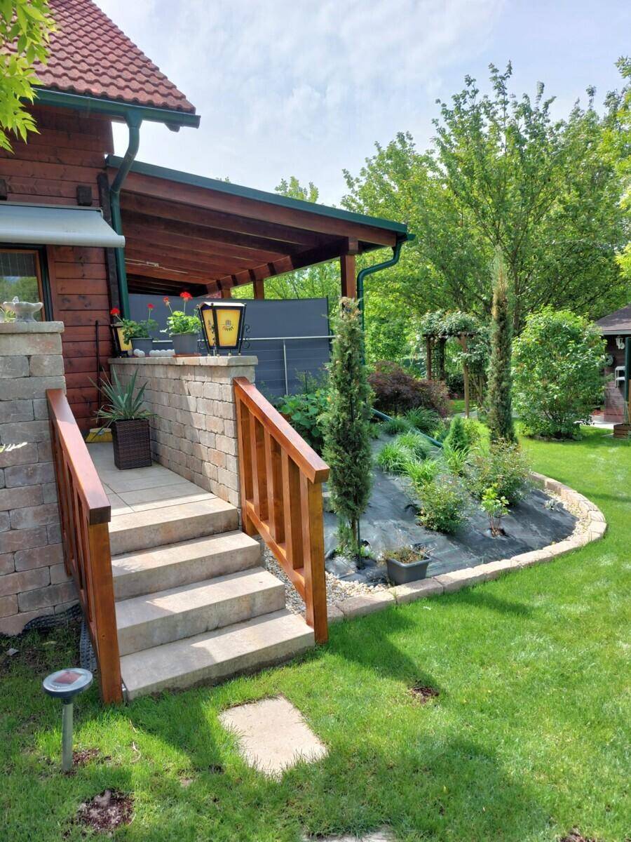 Steintreppe führt zu einer überdachten Terrasse des Holzhauses, umgeben von gepflegten Gartenbeeten.