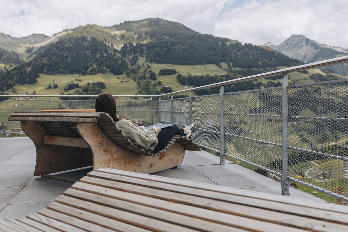 Entspannende Person auf einer Holzliege auf der Terrasse mit atemberaubendem Blick auf die umliegende Berglandschaft und grüne Täler.