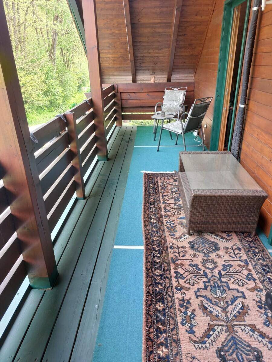 Gemütlicher Balkon mit Holzgeländer, Outdoor-Möbeln und Teppich, mit Blick ins Grüne.