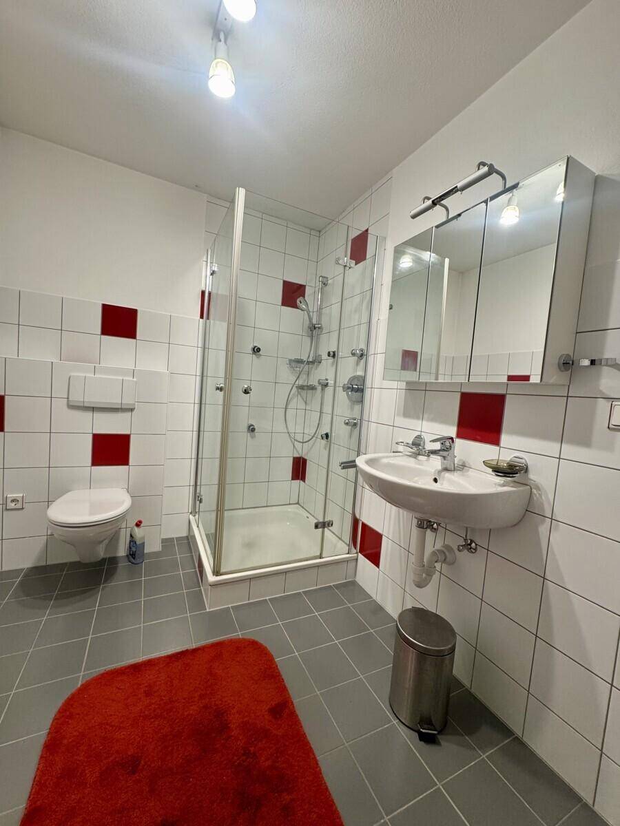 Badezimmer mit Duschkabine, WC und Waschbecken, ausgestattet mit roten Akzentfliesen.