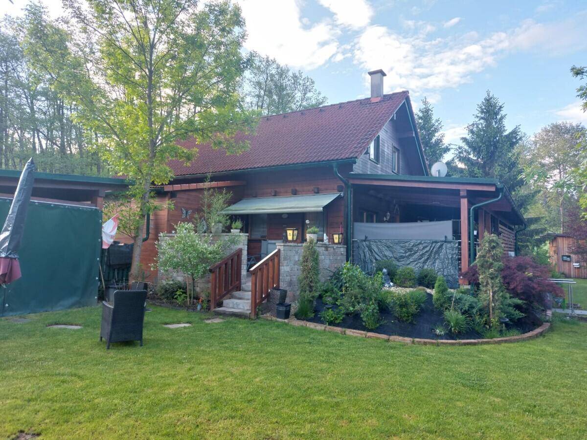Seitlicher Blick auf das Holzhaus mit gepflegtem Rasen, Gartenbeeten und einer überdachten Terrasse.