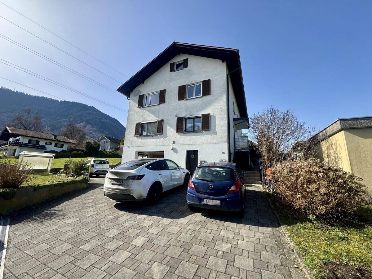 Vorderansicht des Wohnhauses mit gepflasterter Einfahrt, Parkplätzen und Bergblick im Sonnenschein.
