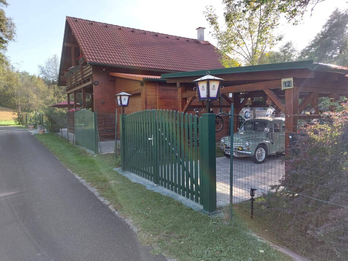 Vorderansicht des Holzhauses mit grünem Zaun und einem Carport, in dem ein Oldtimer parkt.