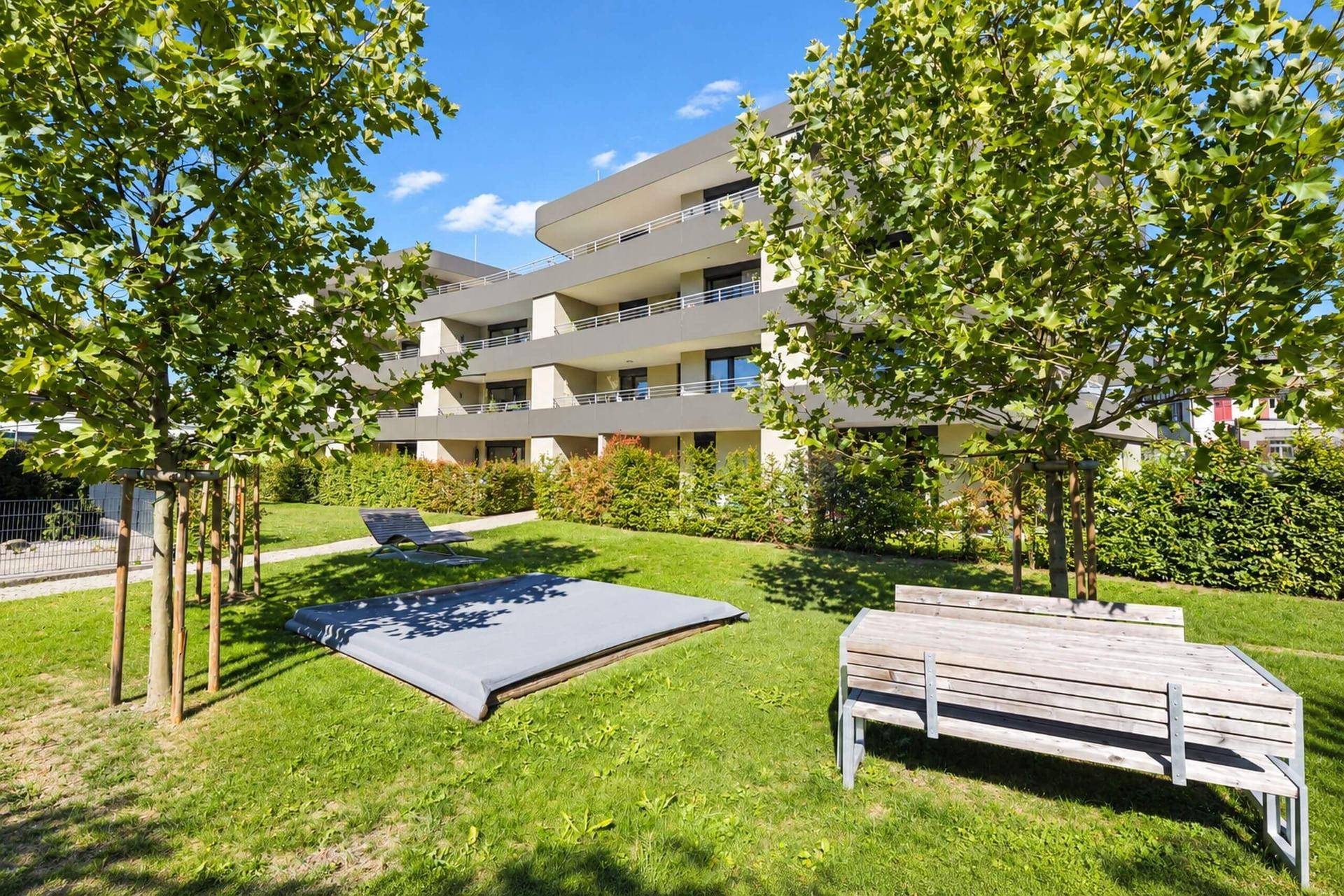 Gepflegte Gartenanlage mit Rasenfläche, Trampolin und Sitzbank vor dem modernen Wohngebäude.