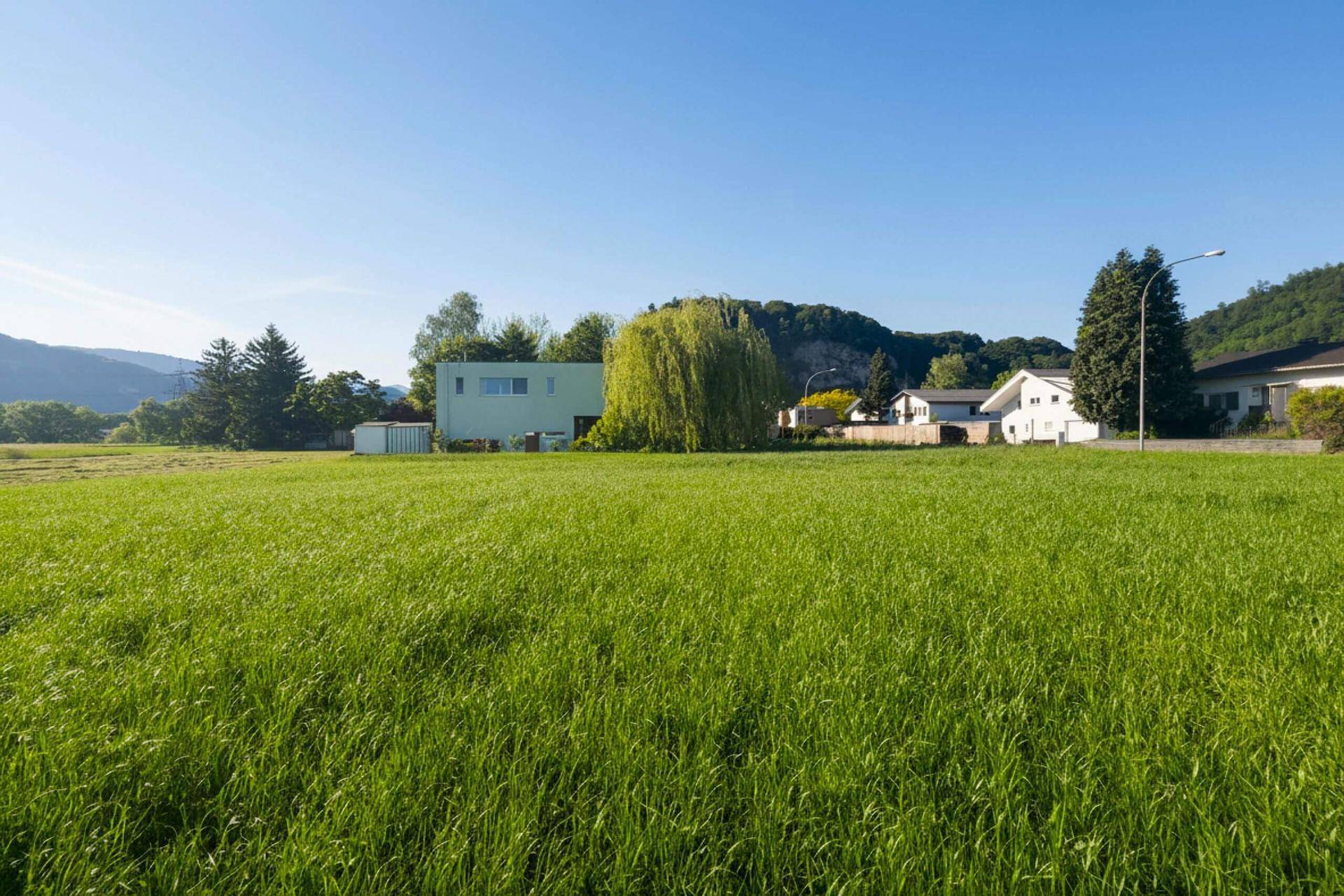 Grünes Feld mit Häusern und Bergen im Hintergrund unter klarem, blauem Himmel.
