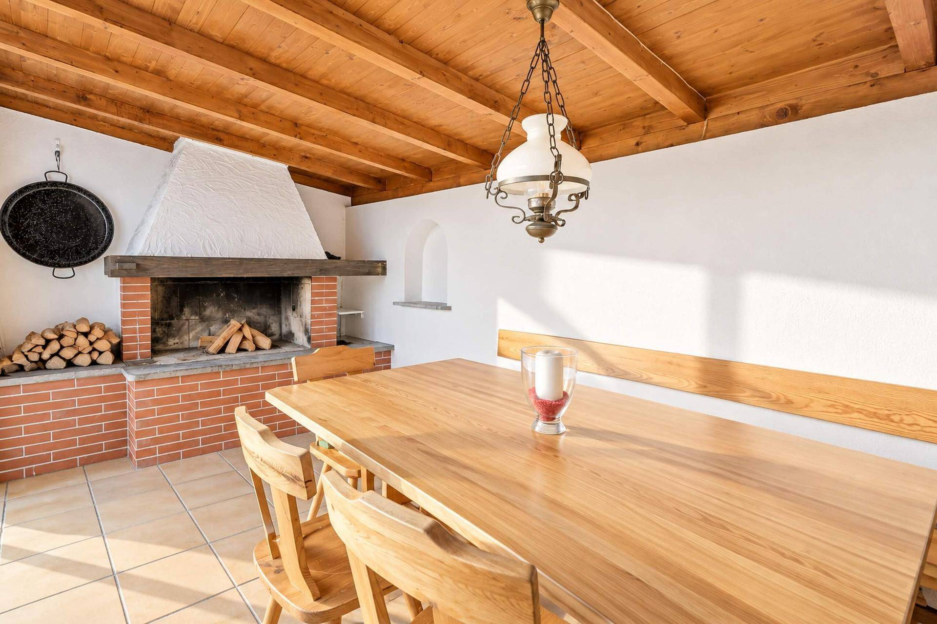 Überdachter Essbereich im Freien mit Holztisch, Bänken und einem gemauerten Kamin.