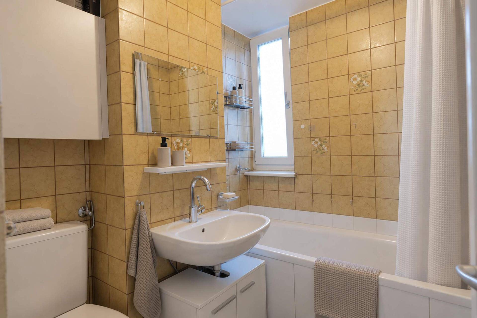 Badezimmer mit Badewanne, Waschbecken, WC und gelben Fliesen im 80er-Jahre-Stil.