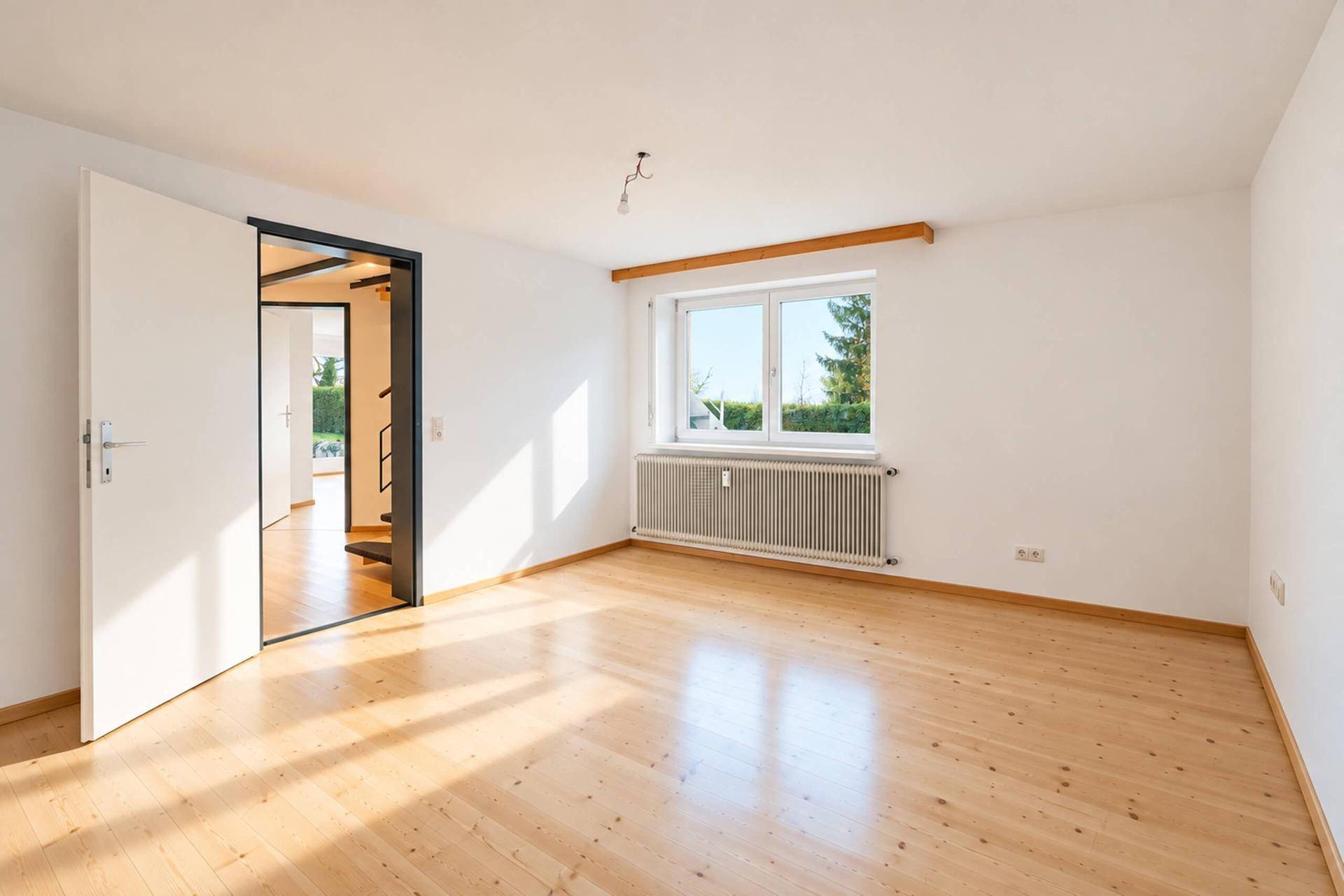 Geräumiges Zimmer mit Holzparkettboden und großem Fenster, das viel Sonnenlicht hereinlässt.