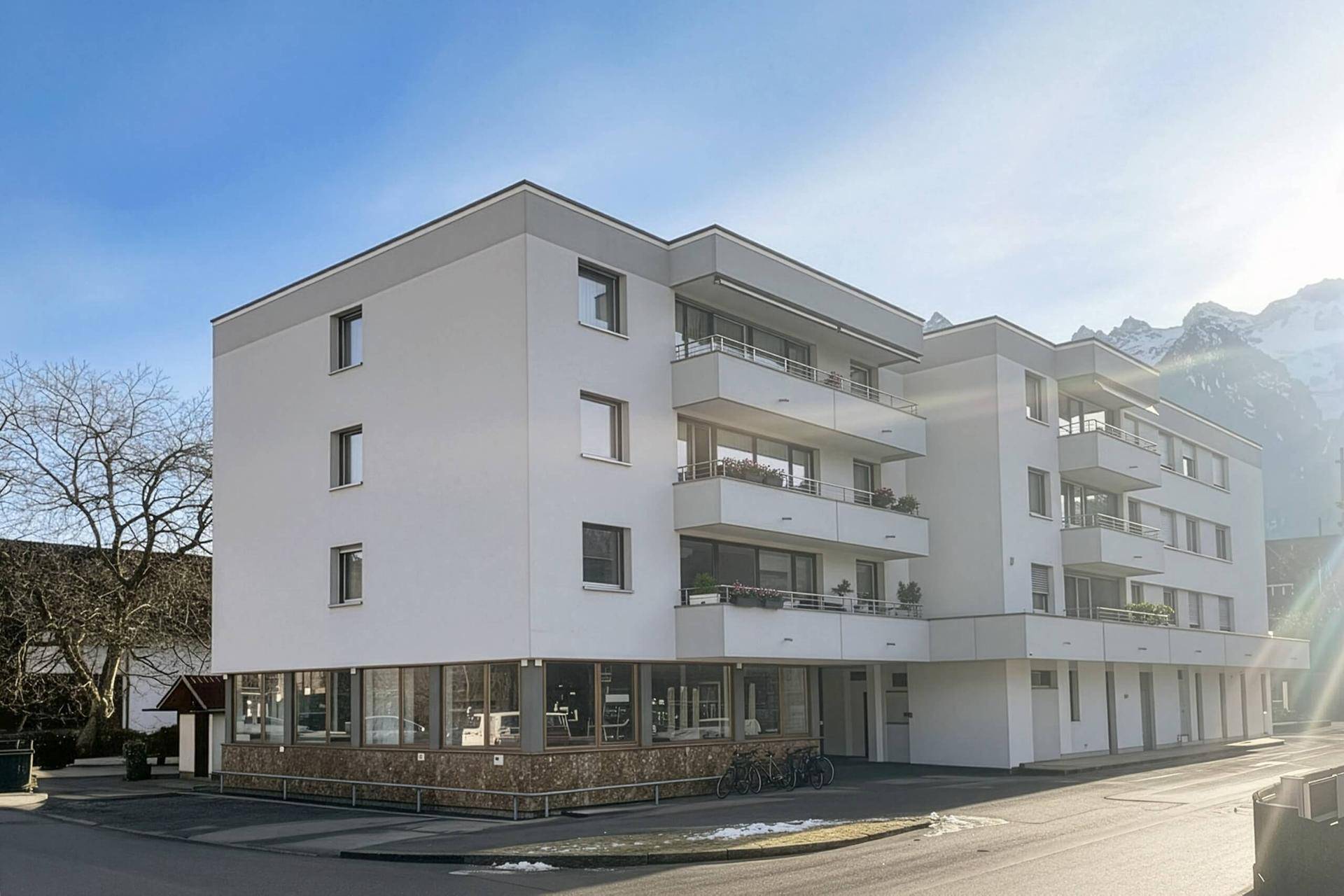 Modernes Wohn- und Geschäftshaus mit Balkonen und großen Fenstern, eingebettet in eine alpine Umgebung.