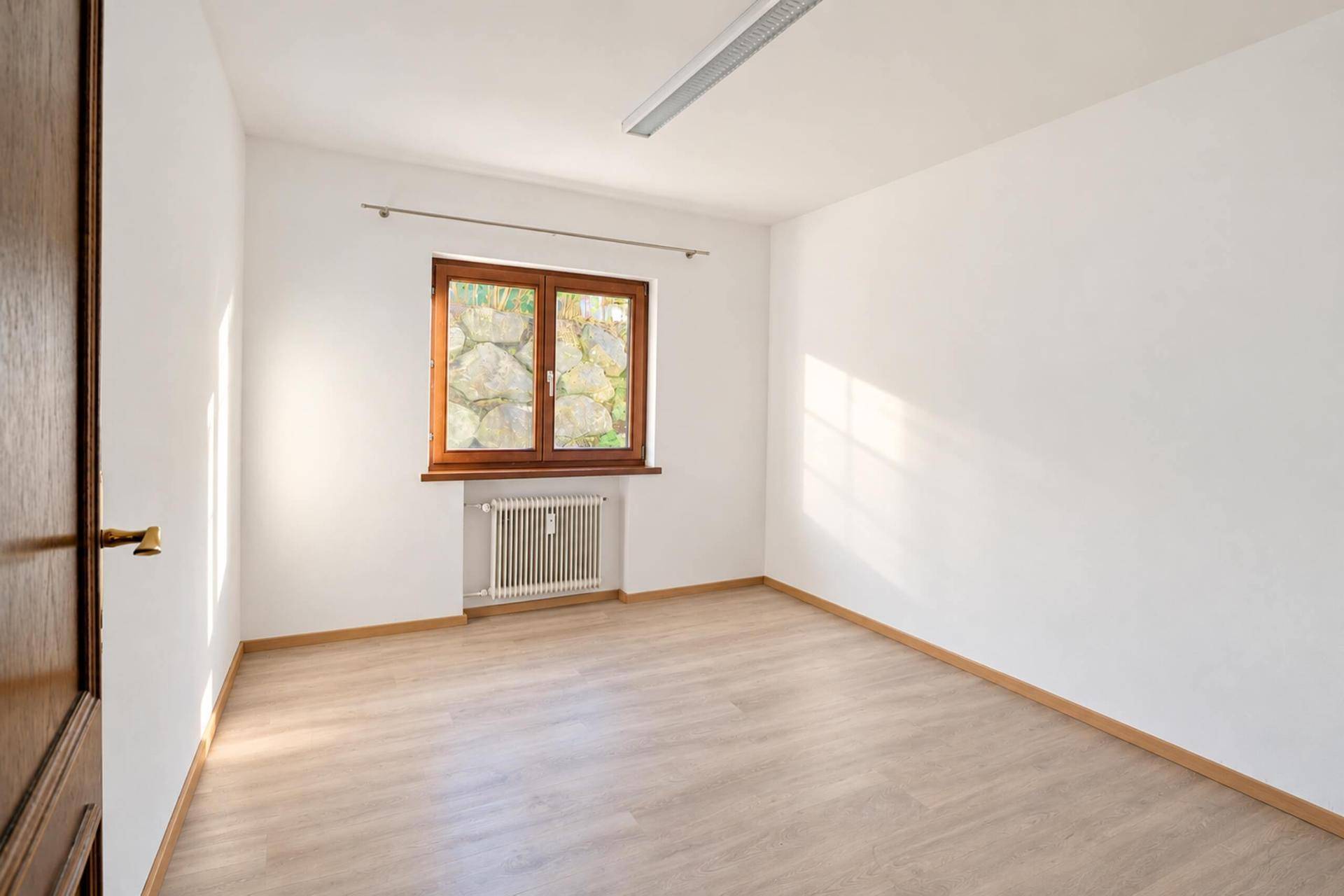 Helles Zimmer mit Laminatboden und Fenster, das einen Blick ins Grüne bietet.