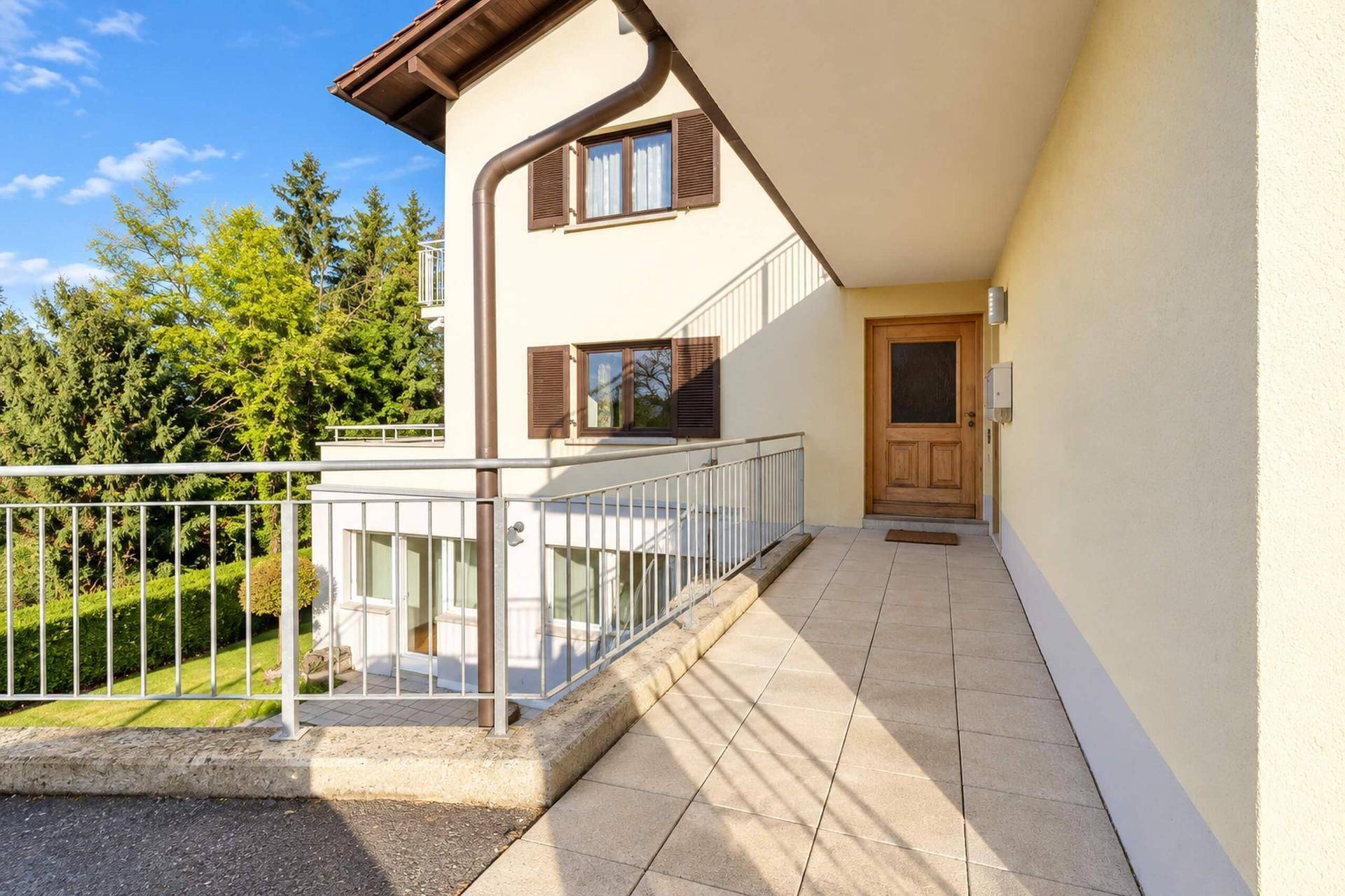 Eingangsbereich des Hauses mit barrierefreier Rampe und Holztür, umgeben von viel Grün.