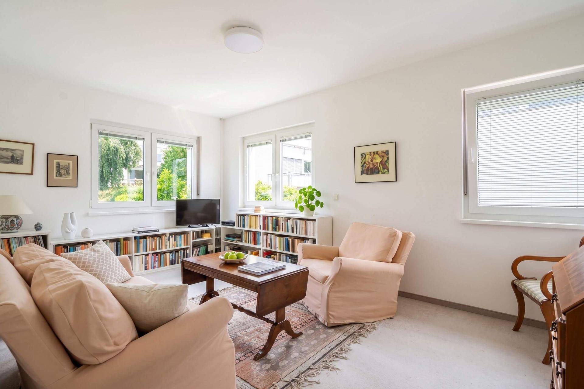 Helles Wohnzimmer mit Teppichboden, beigen Sesseln und Bücherregalen.