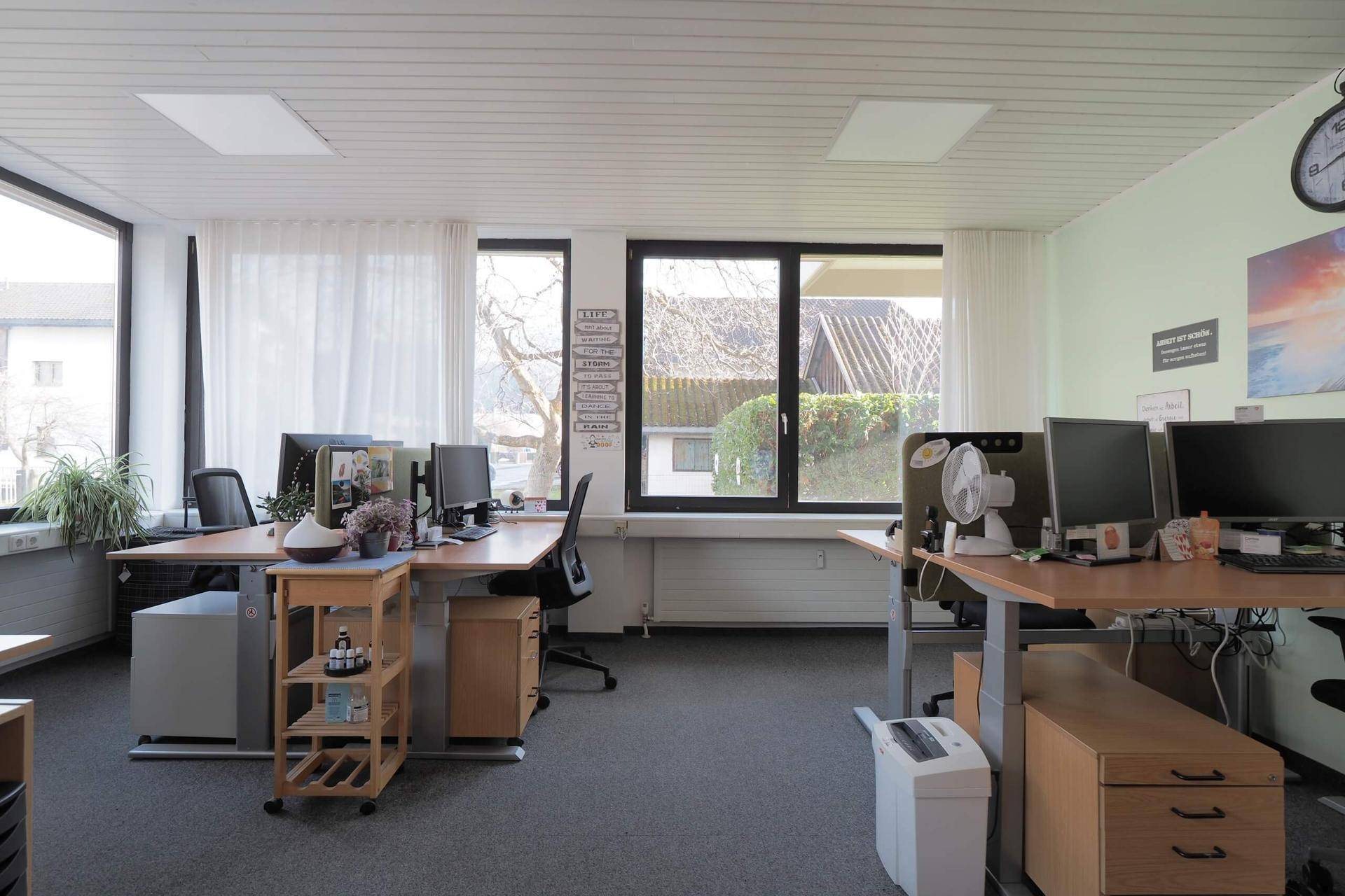 Weiterer Blick in das helle Großraumbüro mit mehreren Schreibtischen, Bürostühlen und großen Fenstern.