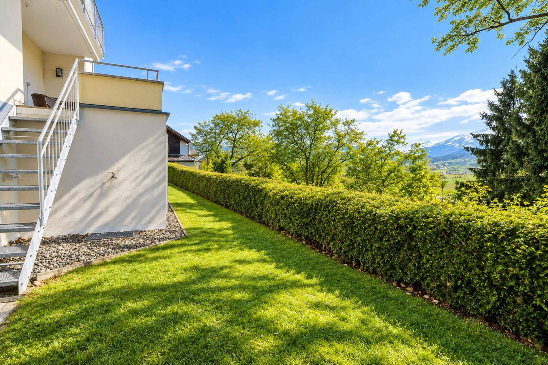 Großer, gepflegter Rasenbereich mit Hecke und Außentreppe, umgeben von grüner Landschaft.