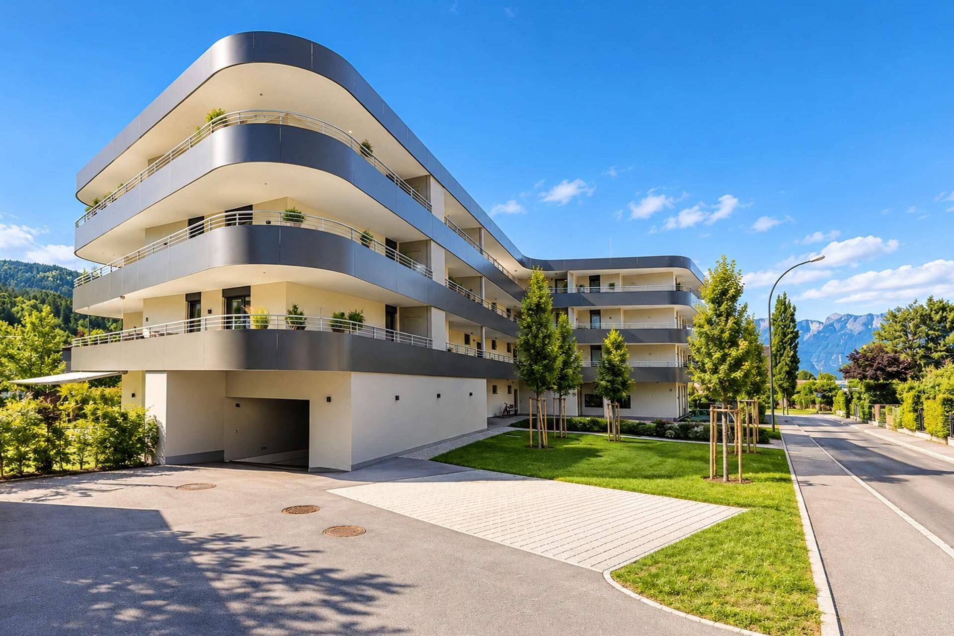 Modernes Wohngebäude mit Tiefgaragenzufahrt, umgeben von Grünflächen und einer Straße mit Bergblick.