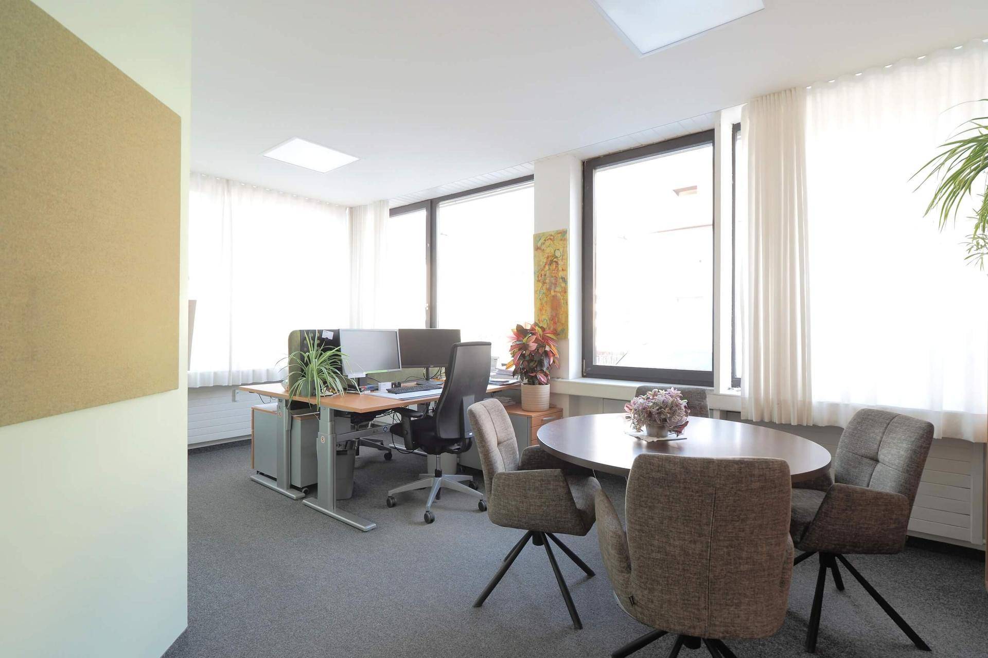 Bürofläche mit einem kleinen Besprechungsbereich und einem Arbeitsplatz im Hintergrund, beleuchtet durch große Fenster.