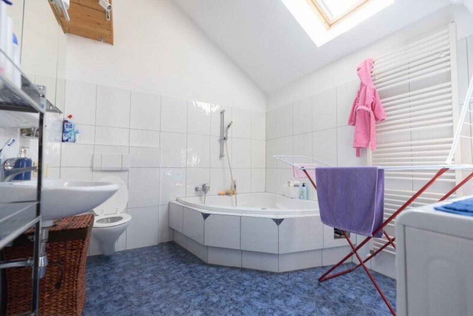 Helles Badezimmer mit Eckbadewanne und weißen Fliesen, ausgestattet mit einem Dachfenster für Tageslicht.