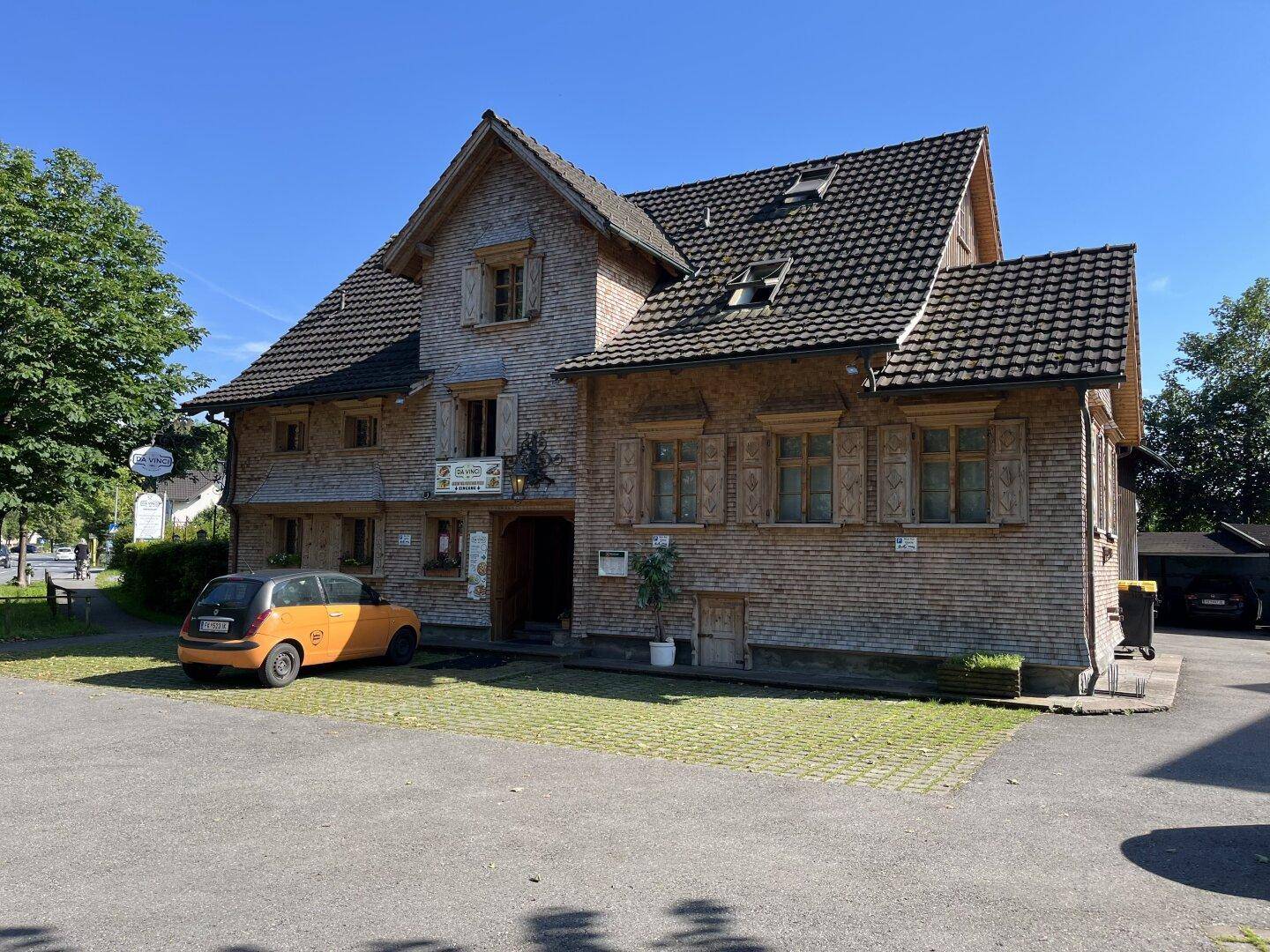 Historisches Gebäude mit Holzschindeln und Satteldach, umgeben von einem gepflasterten Parkplatz und Bäumen.
