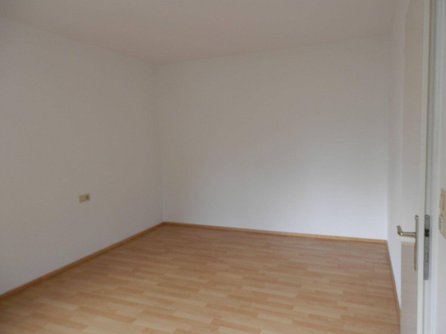 Leeres Zimmer mit hellem Laminatboden und weißen Wänden, bereit zur individuellen Gestaltung.