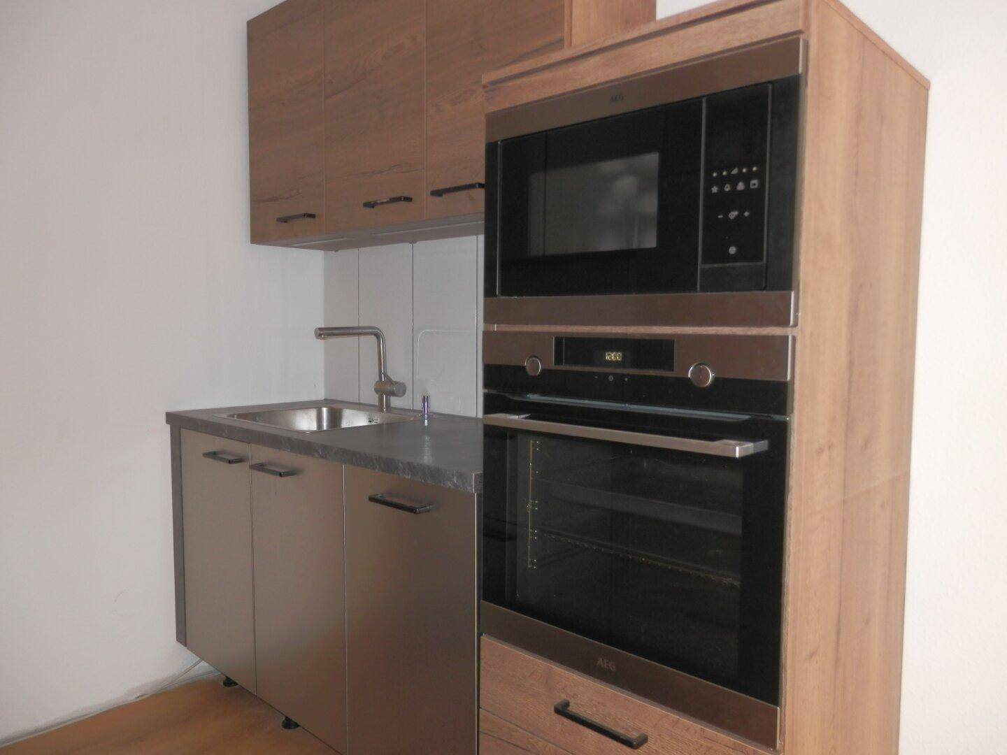 Einbauküche mit Edelstahlspüle, Backofen und Mikrowelle, umgeben von Holzoptik-Schränken.