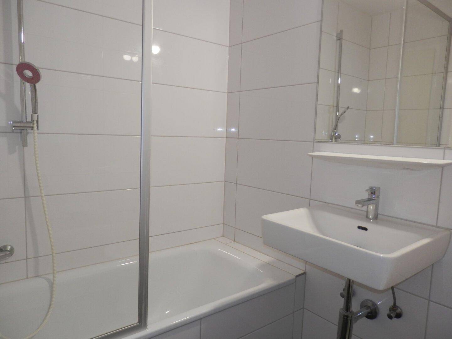 Helles Badezimmer mit weißen Fliesen, Badewanne mit Duschabtrennung und modernem Waschbecken.