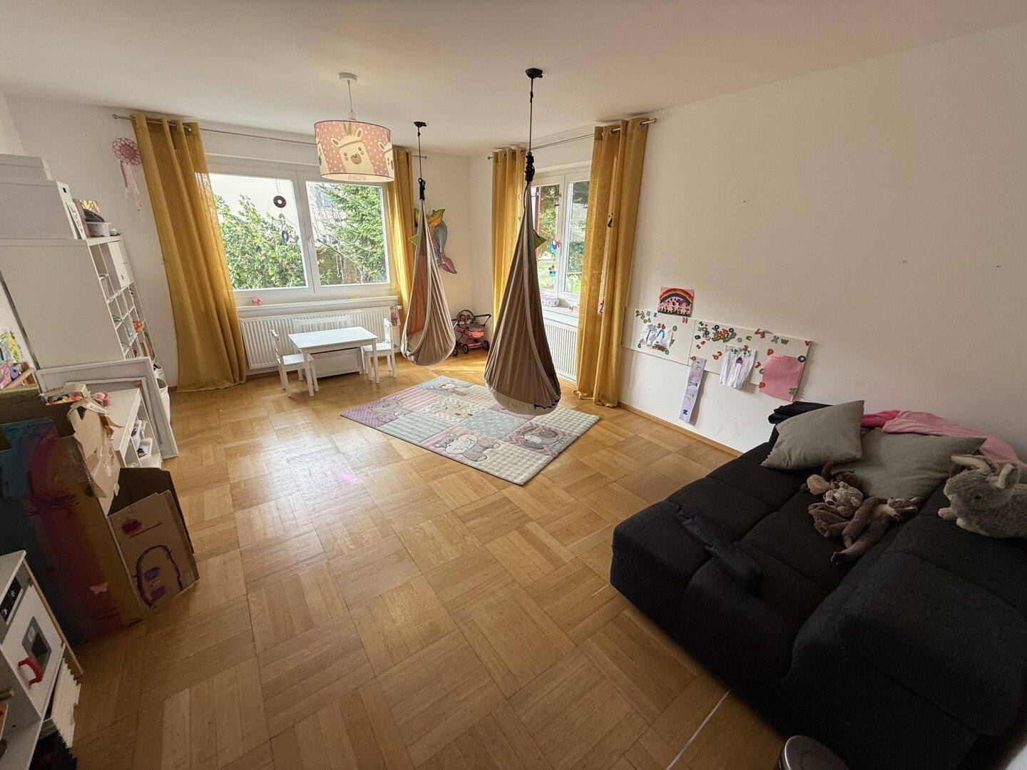 Helles Kinderzimmer mit Parkettboden, großen Fenstern und Spielgeräten.