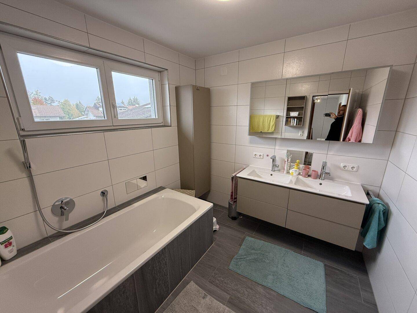 Helles Badezimmer mit Badewanne, Doppelwaschbecken und großem Fenster.