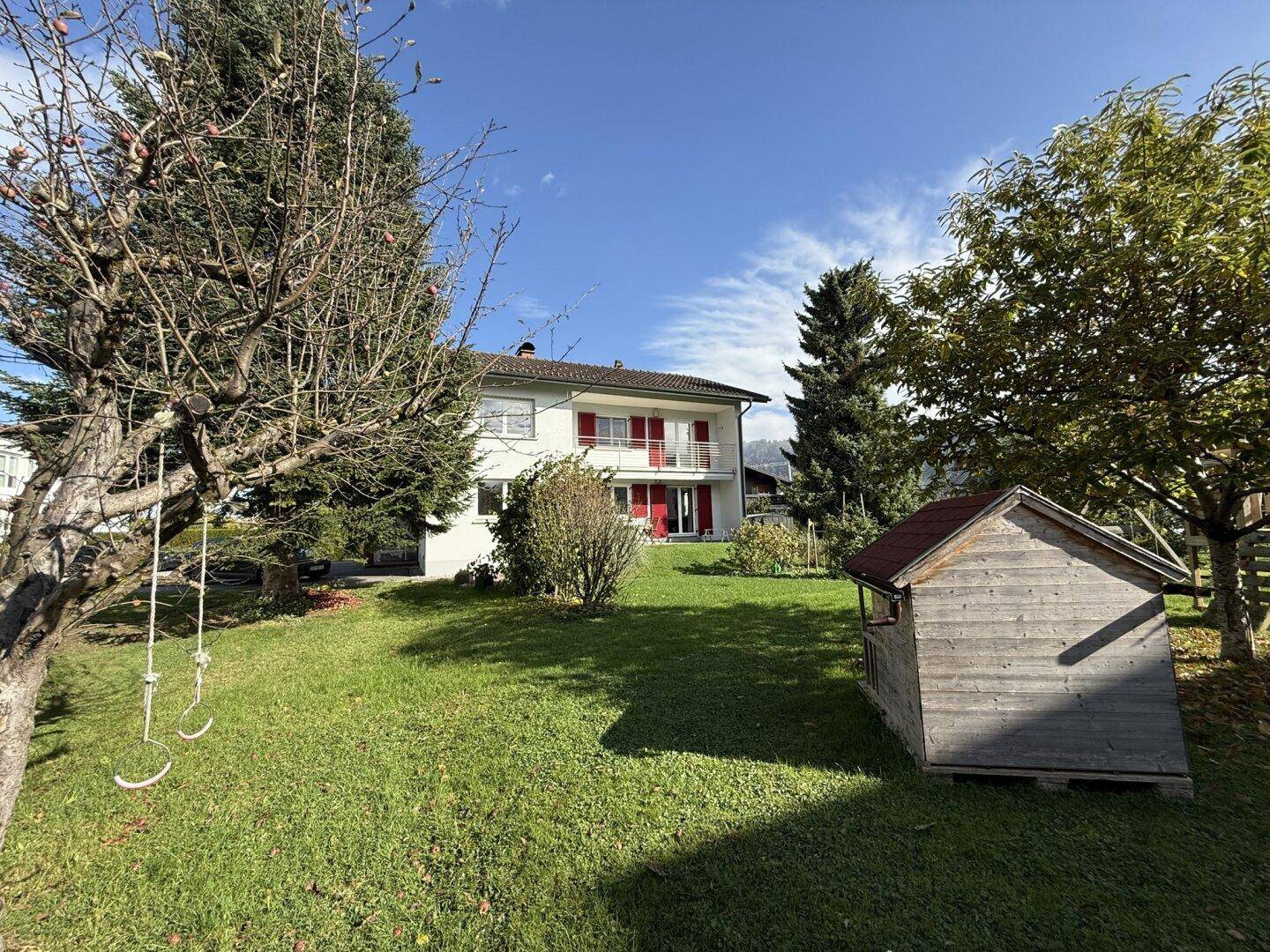 Sonniger Garten mit Rasen, altem Baumbestand und einem kleinen Geräteschuppen im Hintergrund.