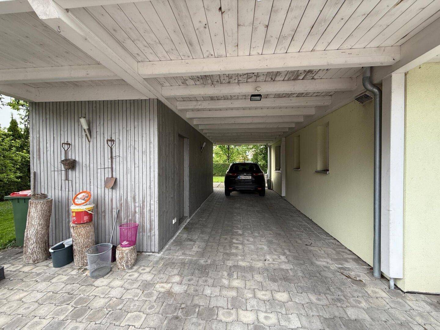 Überdachter Carport mit Holzbalkendecke und gepflastertem Boden für Fahrzeuge.