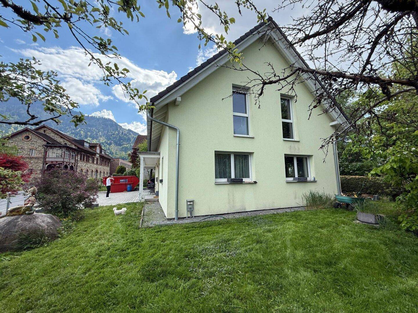 Seitenansicht des Hauses mit heller Fassade, kleinem Rasen und Bergblick.