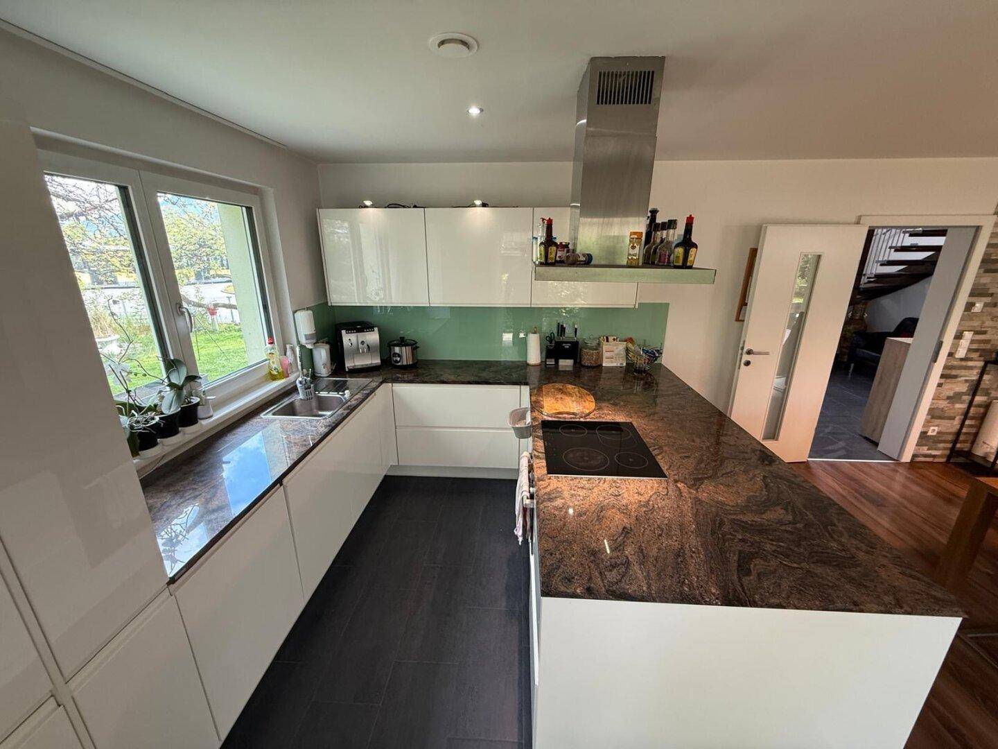 Moderne Küche mit weißen Hochglanzfronten, Fenster zum Garten und dunklem Fliesenboden.