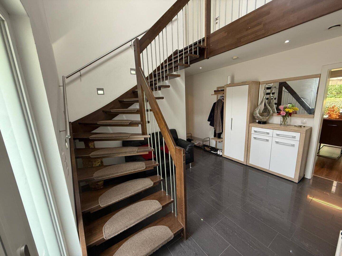 Moderner Eingangsbereich mit offener Holztreppe, dunklem Fliesenboden und Garderobenmöbeln.