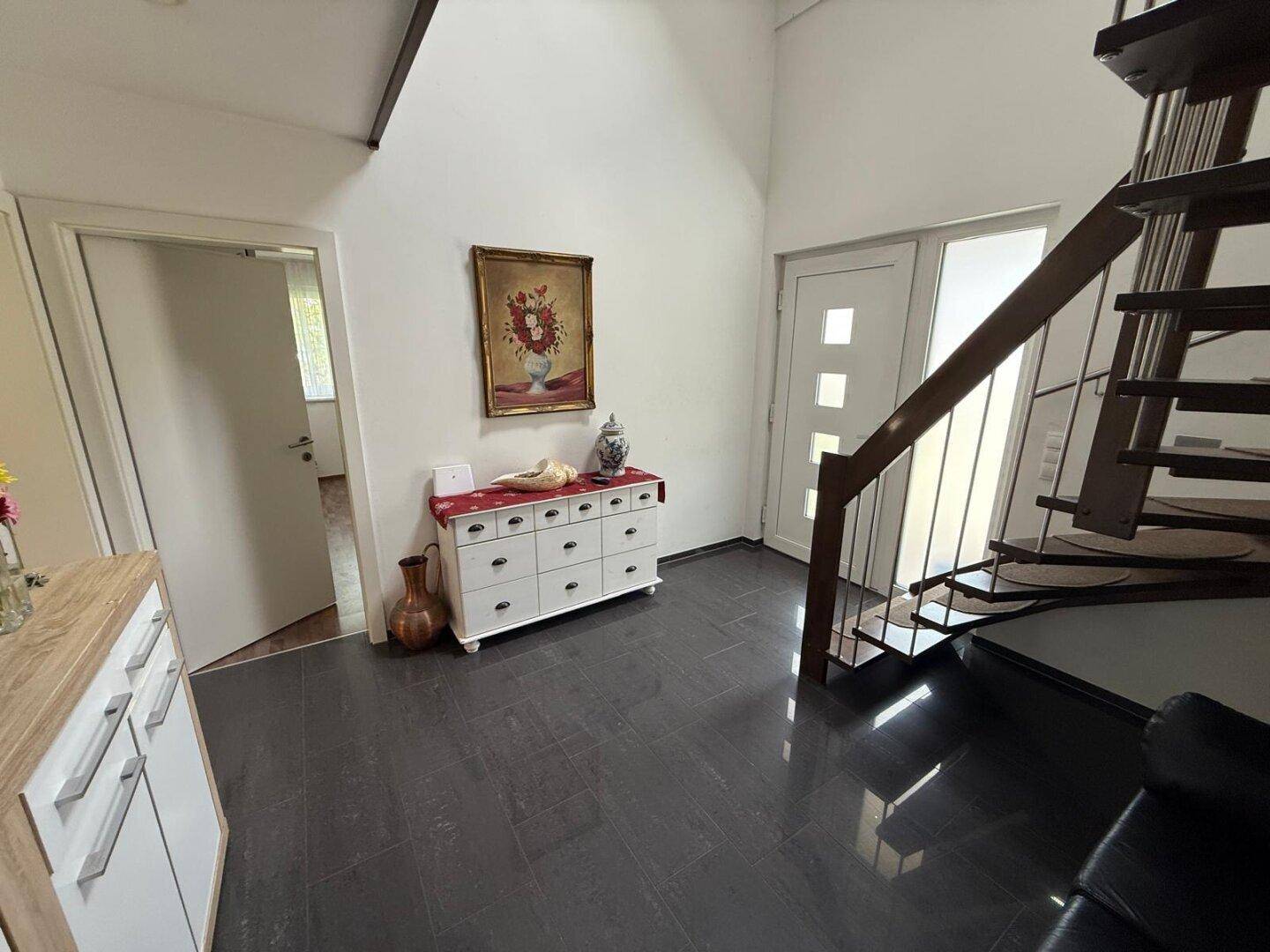 Heller Eingangsbereich mit dunklem Fliesenboden, Holztreppe und weißer Kommode.