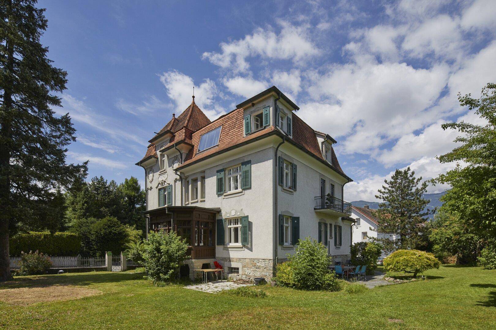 Weitläufiger Gartenbereich mit alter Villa, Baumbestand und Sitzgelegenheiten im Freien unter blauem Himmel.