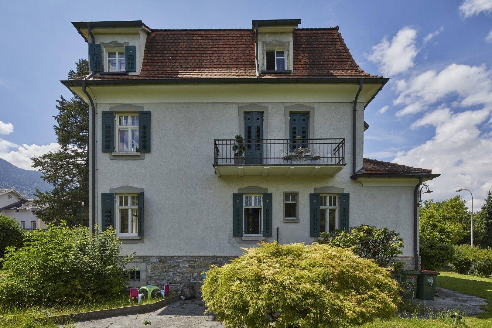 Rückseite der Villa mit kleinem Balkon, grünen Fensterläden und Blick auf den weitläufigen Garten.