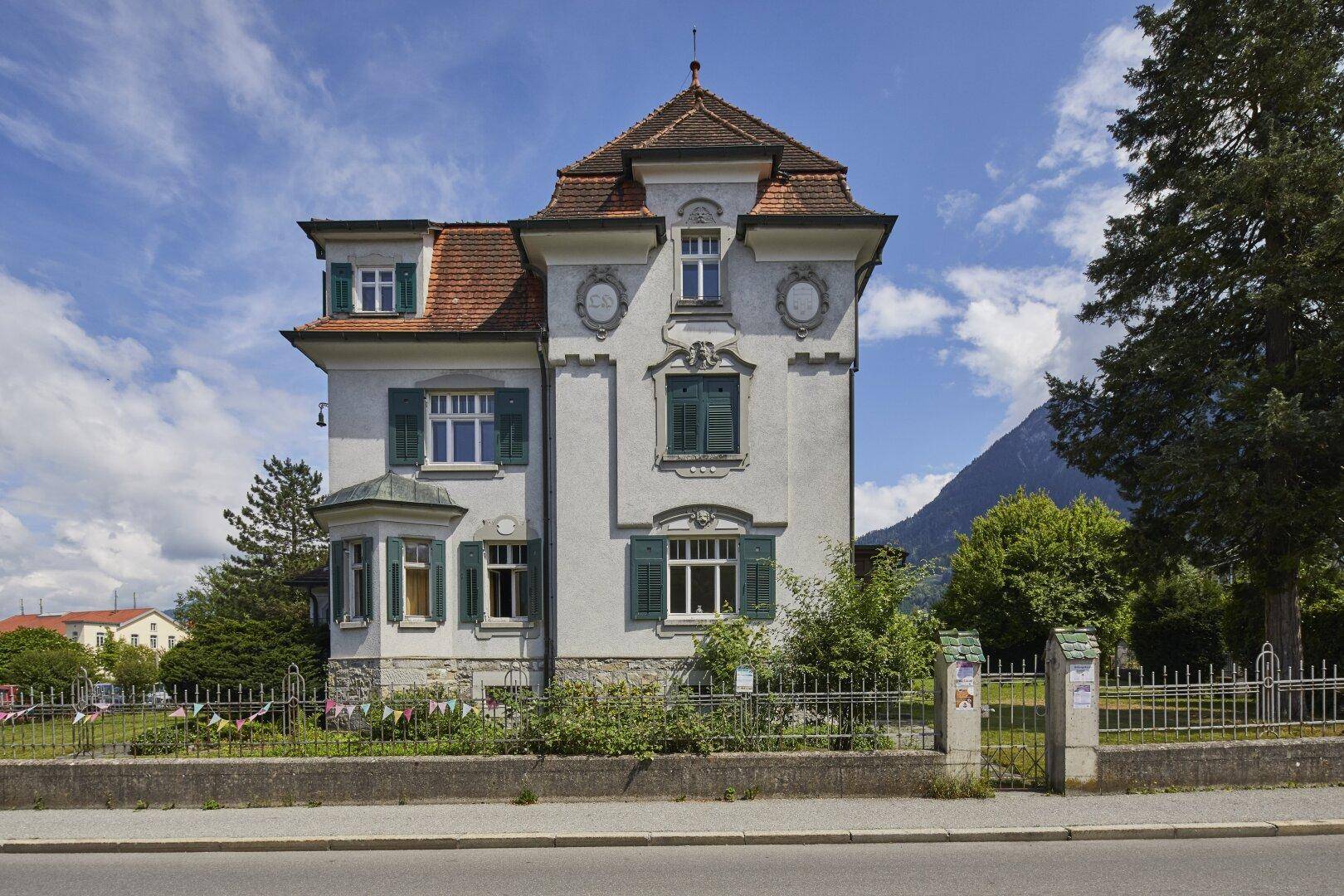 Historische Villa mit eleganter Fassade, grünen Fensterläden und Blick auf die umliegende Berglandschaft.