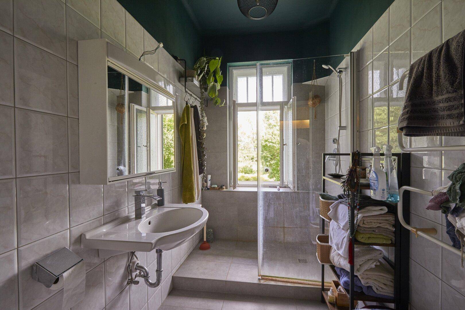 Helles Badezimmer mit Duschkabine, Waschbecken, Fenster und hellen Fliesen an Wänden und Boden.