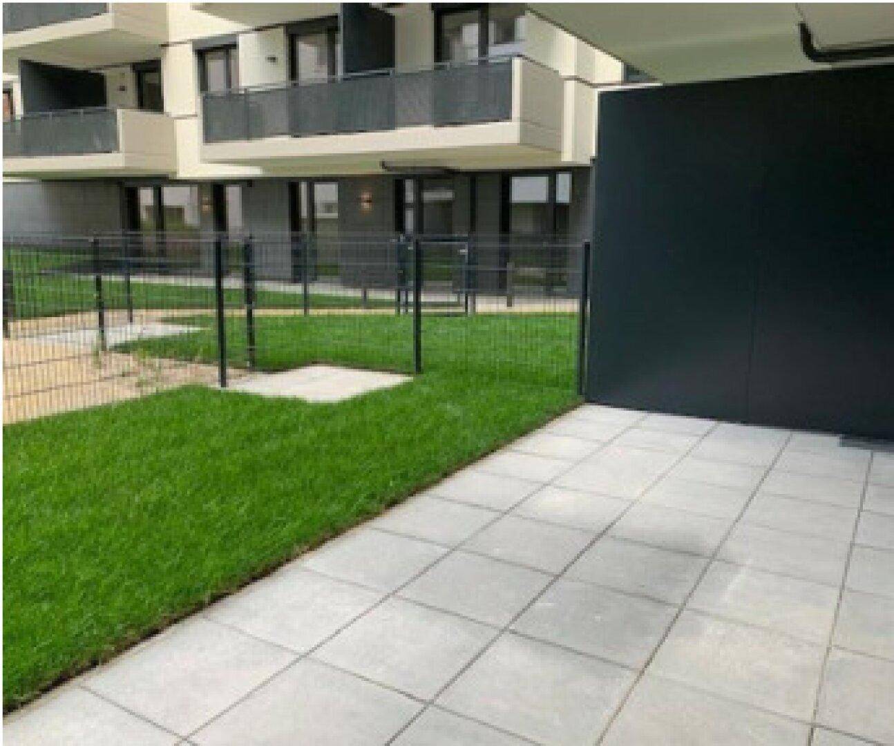 Blick auf den grünen Gartenbereich mit Zaun und die Balkone des modernen Wohngebäudes.