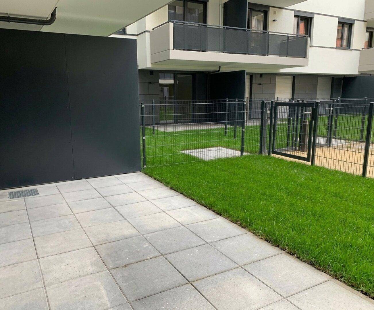 Gepflegter Gartenbereich mit Rasenfläche und Zaun, angrenzend an eine moderne Wohnanlage.