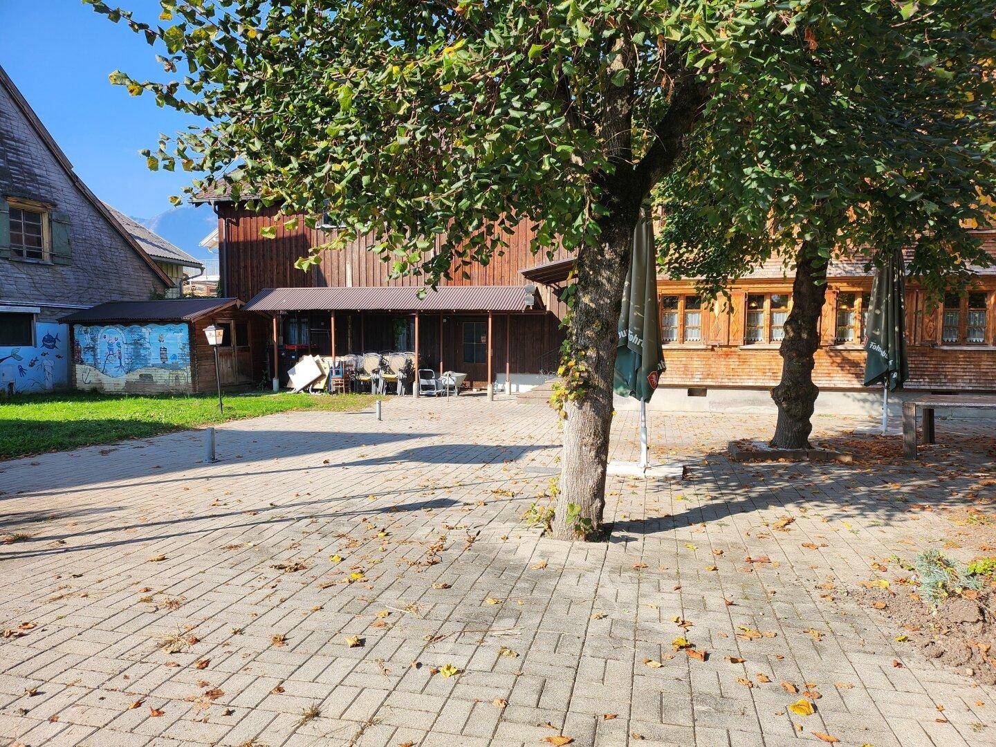 Gepflasterter Außenbereich mit Bäumen und einem traditionellen Gebäude im Hintergrund.