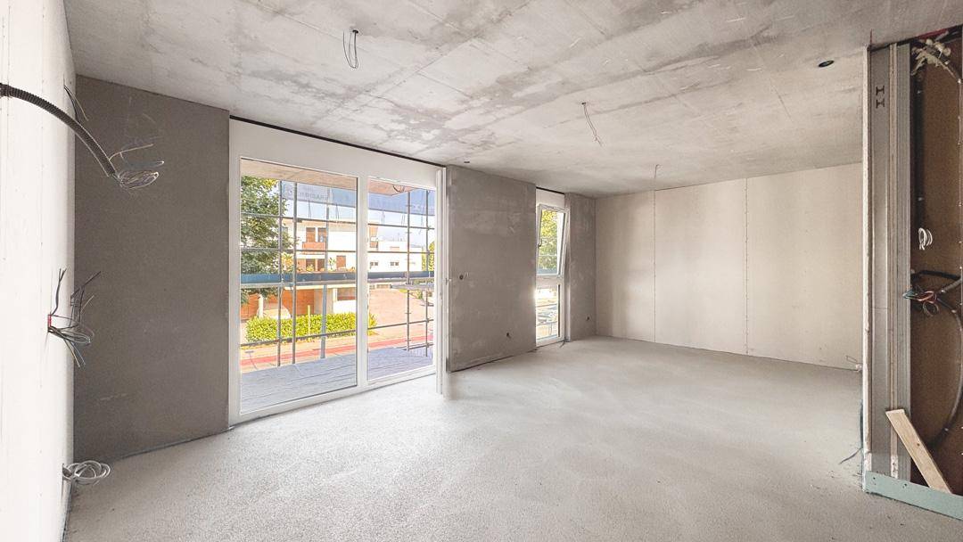 Unfertiger Innenraum im Rohbau mit großen Fenstern, Betonboden und sichtbaren Installationen.