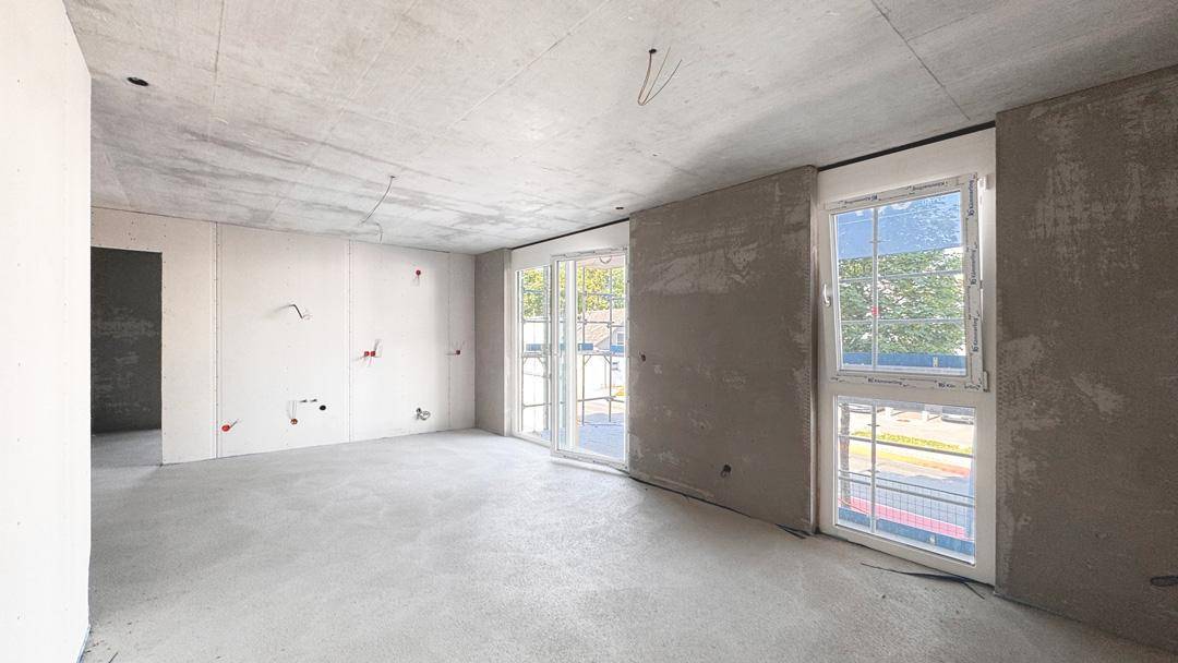 Unfertiger Innenraum im Rohbauzustand mit Betonboden, Decke und sichtbaren Installationen.
