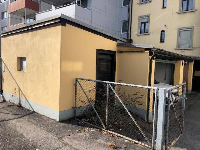 Kleines gelbes Nebengebäude mit einem Garagentor und einem Zaun, das als Parkmöglichkeit dient.